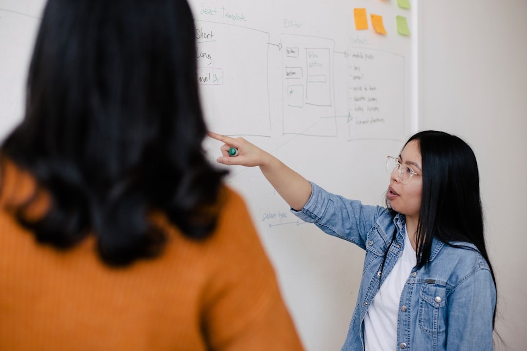 Two women discuss a workflow diagram on a whiteboard.